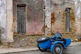 A blue motorcycle with sidecar is parked next to crumbling building.